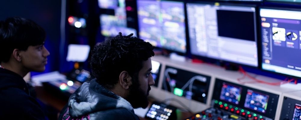 Two students working on broadcast equipment, including various screens and light-up soundboards and switches.