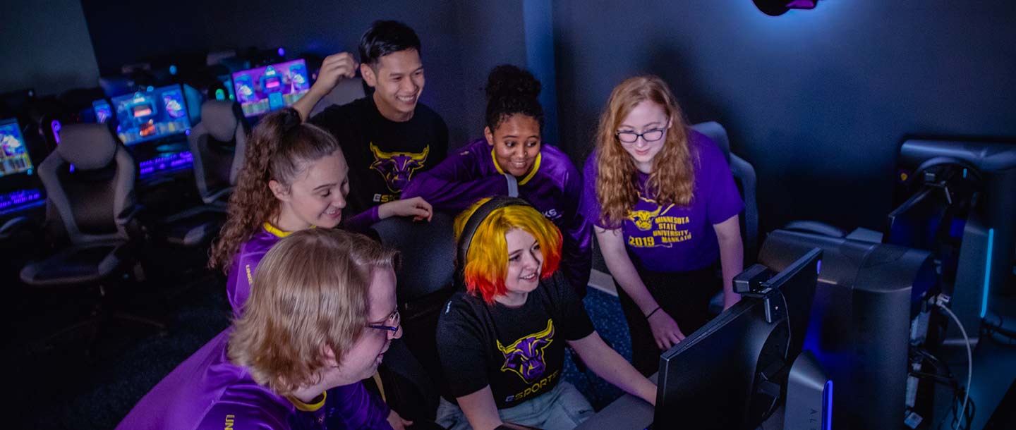 Students in the Esports Training Facility gathering around, cheering, and smiling as a student games on a PC