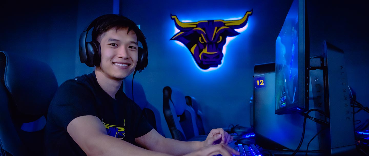 Student wearing headphones, smiling at the camera, and gaming on a PC in the Esports Training Facility