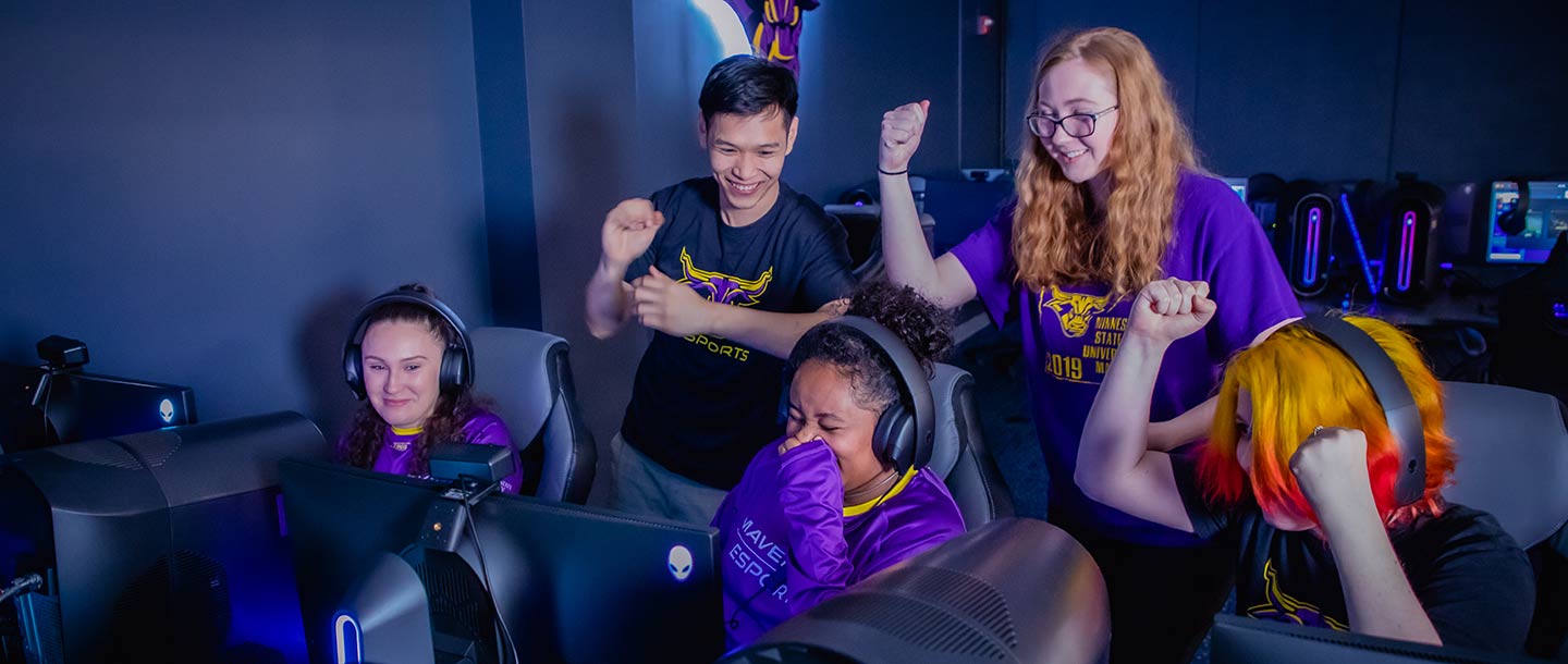 Students gathering around and cheering and smiling as a girl games on a PC in the Esports Training Facility