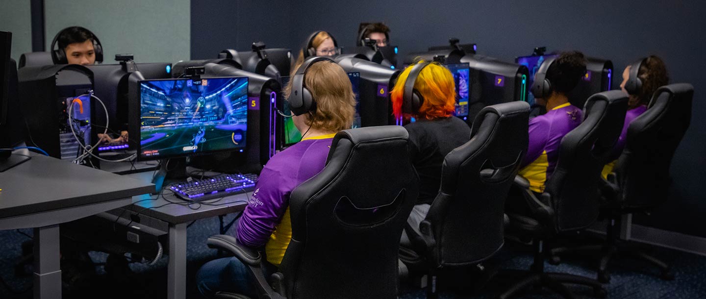 A group of students wearing headphones and purple and gold jerseys gaming on PCs in the Esports Training Facility