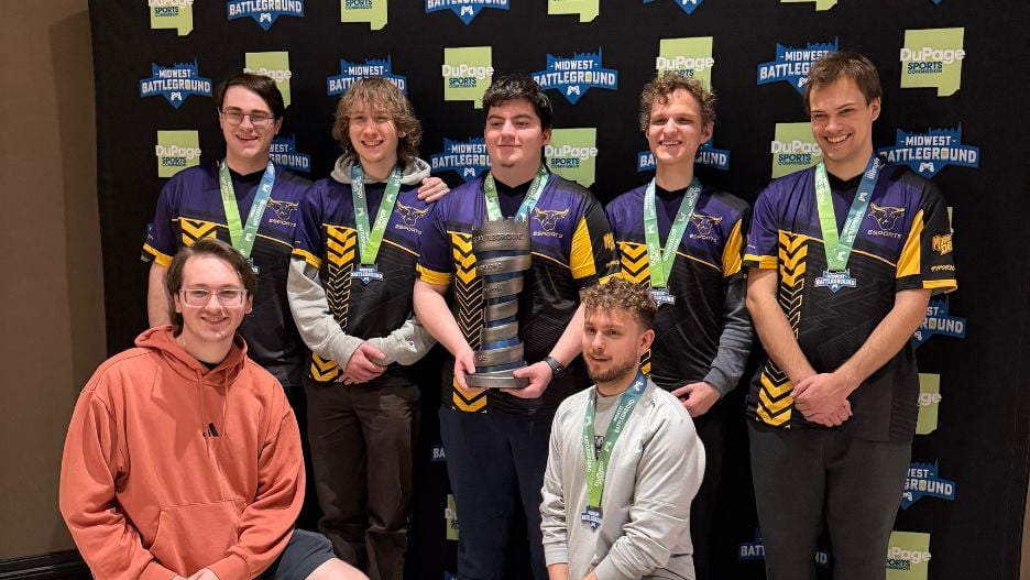 A team of varsity esports students wearing Mavericks jerseys posing with their coaches and holding a trophy they won at Midwest Battleground LAN