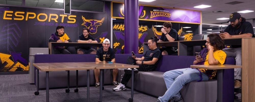 A group of students hanging out in the gaming lounge, sitting on couches and high chairs, chatting, smiling, and using their phones.