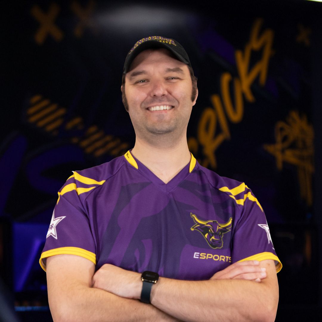 Mitch Wallerstedt, smiling with arms folded, wearing a baseball cap and purple and gold esports jersey
