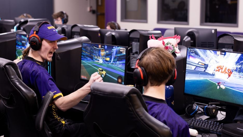 Two students gaming at the open play gaming stations in the esports facility, cheering and smiling