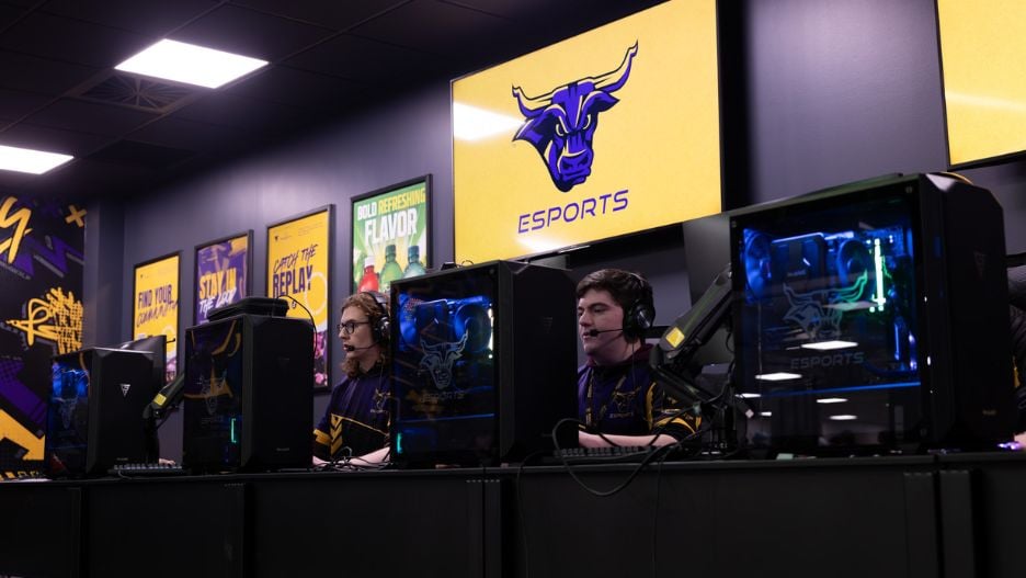 Two students wearing headsets and Maverick Esports jerseys gaming on PCs in the esports facility, with colorful purple and gold graphics and murals on the wall.