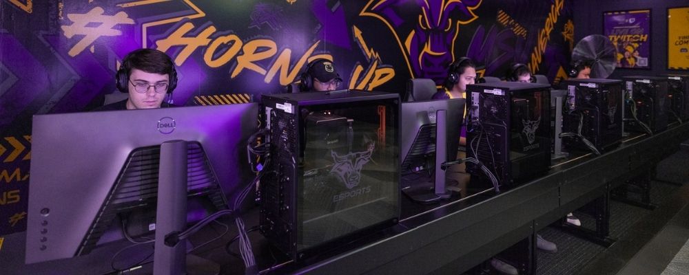 A row of varsity esports players wearing headphones and concentrating on practicing their gaming skills on computers in the training facility, with a purple and gold Mavericks branded mural on the wall in the background.